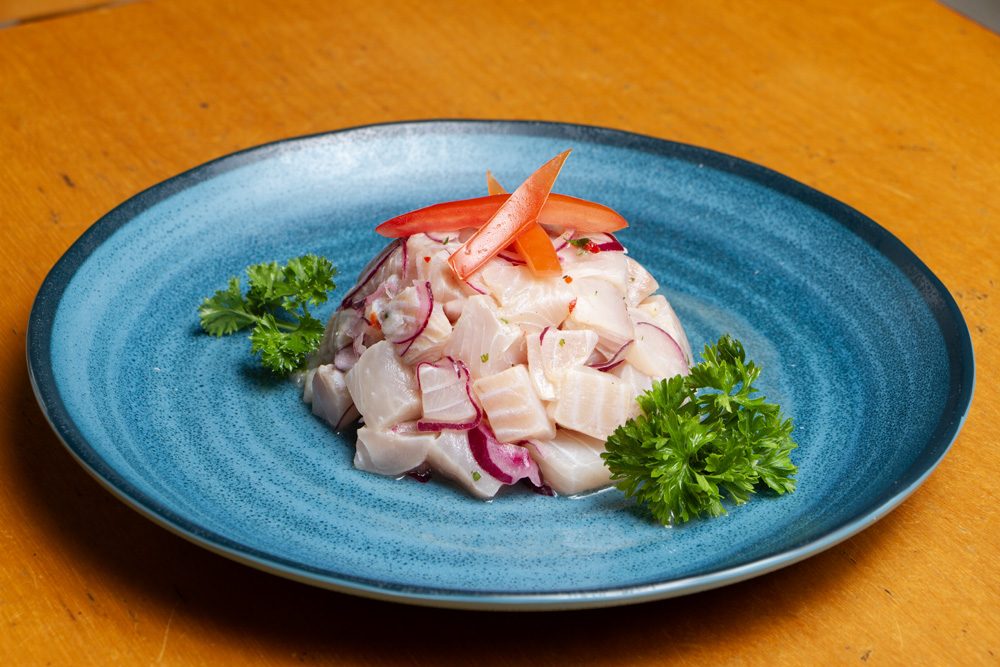 Ceviche de peixe branco em cubos com cebola roxa e tomate, servido em prato azul sobre mesa de madeira